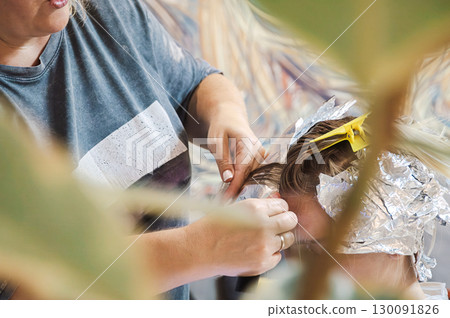 Hairdresser woman applying foil to teen boy's hair for highlights coloring. Professional hair stylist working on beauty treatment for customer. Hairdresser woman applying foil to teen boy's hair for highlights coloring. Professional hair stylist working on beauty treatment for customer. 130091826