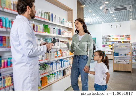 Young mom with a cute kid choosing medicine in the pharmacy, male pharmacist helping her 130091917