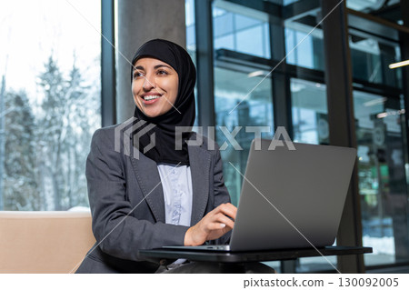 Young woman in hijab working on laptop and smiling 130092005