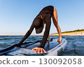 Young adult slim woman perform Yoga Downward-Facing Dog surfing SUP paddleboard Baltic Sea waves warm sunset sun lights summer day. Sunlight sea ocean waves enhance harmony body, strength mindfulness 130092058
