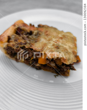 Slice of delicious homemade meat pie with melted cheese and vegetables on white plate 130092384