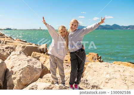 Happy children sisters posing on rocks near sea on sunny rocky coastline day with raised arms Happy children sisters posing on rocks near sea on sunny rocky coastline day with raised arms 130092586
