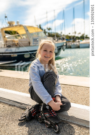 Child sitting in rollerblades on harbor curb with white yachts in background 130092601