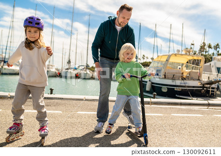Parent supporting blond son riding scooter on dockside near yachts with skating daughter 130092611
