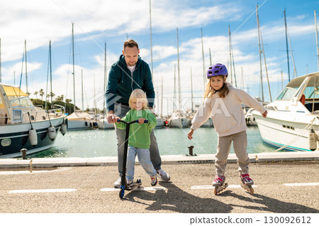 Smiling man supporting cheerful blond boy on scooter along waterfront pier with skating daughter Smiling man supporting cheerful blond boy on scooter along waterfront pier with skating daughter 130092612