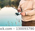 A young Caucasian man with short hair is fishing by a calm lake. He holds a fishing rod and reel,  130093790