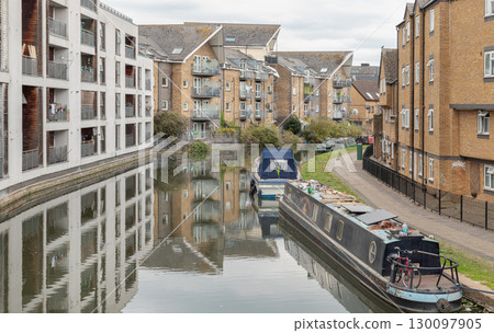 a tranquil scene along the Grand Union Canal, featuring residential buildings lining the canal bank with narrowboats moored in the water. a tranquil scene along the Grand Union Canal, featuring residential buildings lining the canal bank with narrowboats moored in the water. 130097905