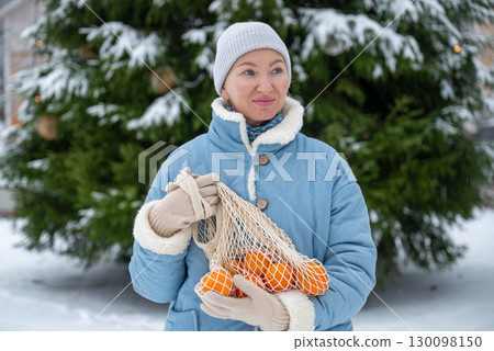 Woman in blue winter coat holding mesh bag with tangerines outdoors in snow 130098150