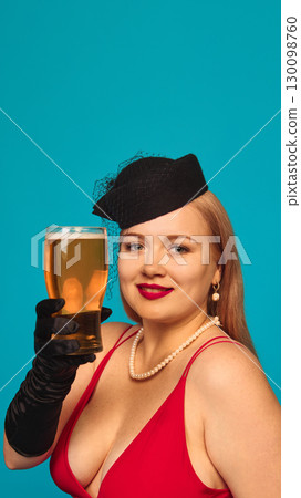 Woman in red dress with pearls and hat holding beer glass with smile on blue background 130098760