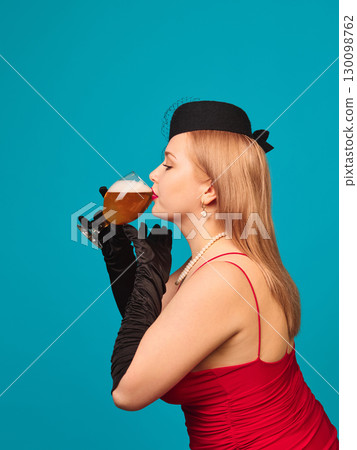 Woman in red dress with pearl necklace drinking beer glass in profile on blue background 130098762