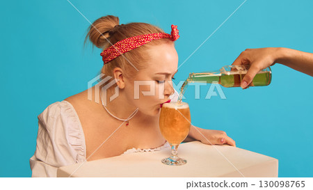 Woman drinking beer from glass while bottle pouring on blue background 130098765
