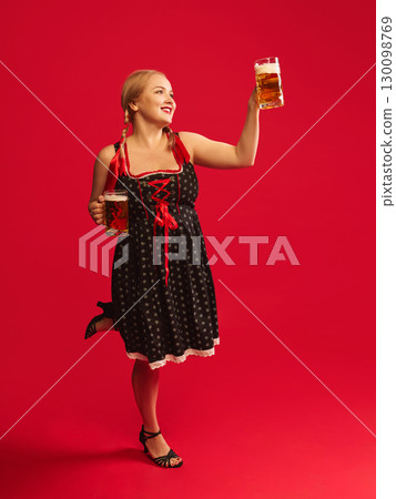 Happy woman in Oktoberfest dress raising two beer mugs with joyful smile against red background Happy woman in Oktoberfest dress raising two beer mugs with joyful smile against red background 130098769