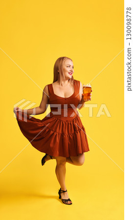 Smiling woman in red dress holding glass of beer with playful joy on yellow background Smiling woman in red dress holding glass of beer with playful joy on yellow background 130098778