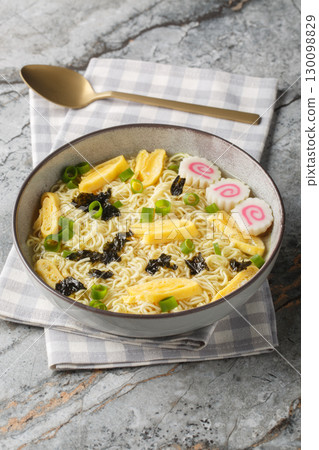 Saimin is a Hawaiian noodle soup consisting of soft wheat egg noodles served in a hot dashi garnished with diced green onions, kamaboko and omelette closeup in the plate. Vertical 130098829