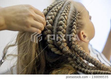 Skilled hands work meticulously to create stunning cornrow braids, enhancing the girl's hairstyle in a chic salon atmosphere. Attention to detail shines as creativity unfolds. Skilled hands work meticulously to create stunning cornrow braids, enhancing the girl's hairstyle in a chic salon atmosphere. Attention to detail shines as creativity unfolds. 130099535
