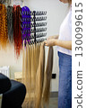 A stylist carefully selects a long strand of golden hair extensions while colorful options hang above. This lively salon showcases a variety of hair textures and colors, inviting creativity. 130099615