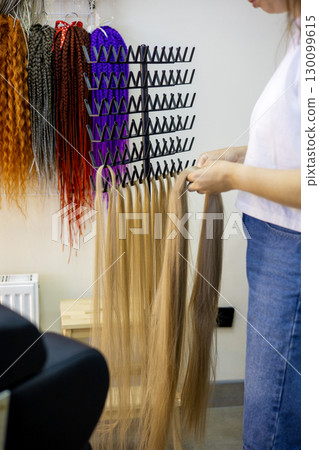 A stylist carefully selects a long strand of golden hair extensions while colorful options hang above. This lively salon showcases a variety of hair textures and colors, inviting creativity. 130099615