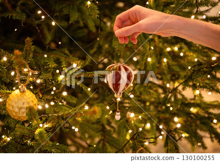 Hand hanging ornament on decorated Christmas tree with glowing lights 130100255