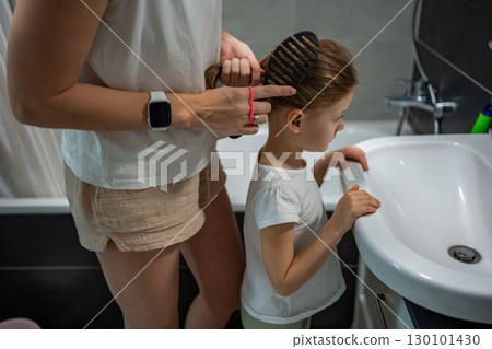 Mother braiding her daughter hair in the bathroom before going to school or kindergarten. Concept of morning preparation, care and family routine in childhood. 130101430