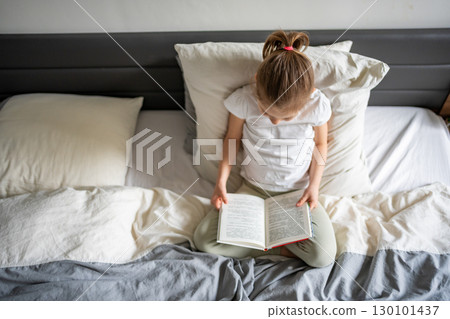 Little girl sitting on her parents bed and reading a book in the bedroom. Concept of quiet time, concentration and love for reading in early childhood. 130101437