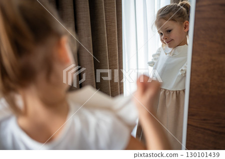 Little girl standing in front of a mirror and holding a dress against herself. Concept of self-expression, imagination and early interest in clothing and appearance. 130101439
