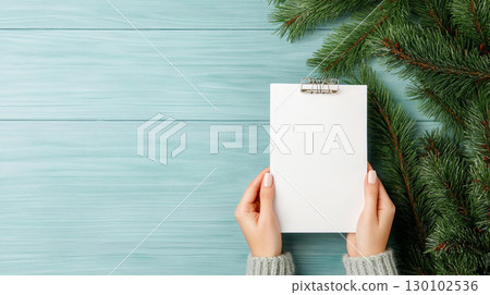 person holding blank notepad surrounded by pine branches on blue wooden surface, evoking cozy winter atmosphere 130102536