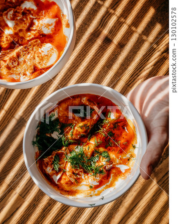 Hand gripping wooden bowl filled with traditional turkish shakshuka, garnished with yogurt, tomato sauce, spices, dill, beside companion bowl under soft natural illumination. Bowl of turkish shakshuka 130102578