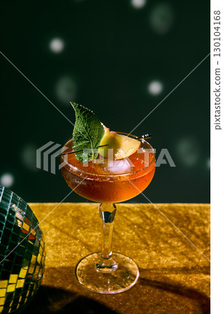 Red cocktail with mint leaf and fruit garnish on glitter table with disco vibe 130104168
