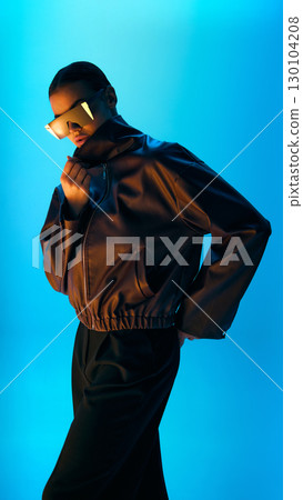 Model in oversized leather jacket and futuristic sunglasses posing on blue neon light background 130104208
