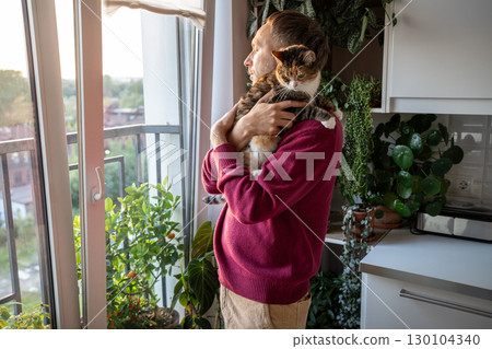 Thoughtful man holding cat while gazing out window on quiet day off in peaceful solitude, slow life 130104340