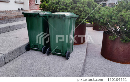 Two green trash bins placed beside large planters in a clean urban setting 130104531