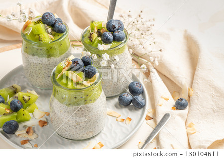 Healthy breakfast chia pudding with kiwi, blueberries and coconut slices, three portions in glass jars on a white table. 130104611