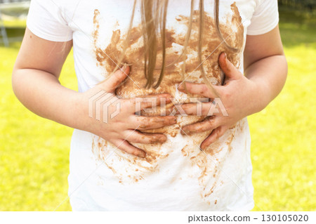 Girl child staining her white t-shirt with stain while enjoying a delicious chocolate bar in garden Girl child staining her white t-shirt with stain while enjoying a delicious chocolate bar in garden 130105020