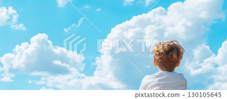 Serene image of a young boy standing in a natural setting gazing up at the picturesque sky filled with fluffy white clouds  The child appears to be lost in thought 130105645