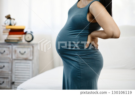 Side view of pregnant woman in blue dress holding her lower back with hand showing discomfort in bedroom Side view of pregnant woman in blue dress holding her lower back with hand showing discomfort in bedroom 130106119