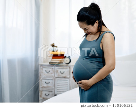 Asian Pregnant woman in blue dress standing indoors holding belly with both hands looking down lovingly in bedroom 130106121