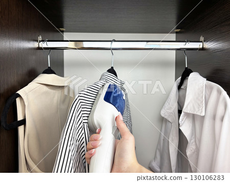 Woman ironing clothes with steam iron at wardrobe. Clothes care and garment steaming. Fashion, laundry, household chores, wrinkle removal. Business and casual clothing care 130106283
