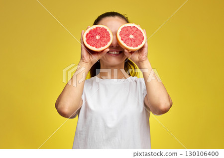 Caucasian woman covering her eye with grapefruit on yellow background 130106400