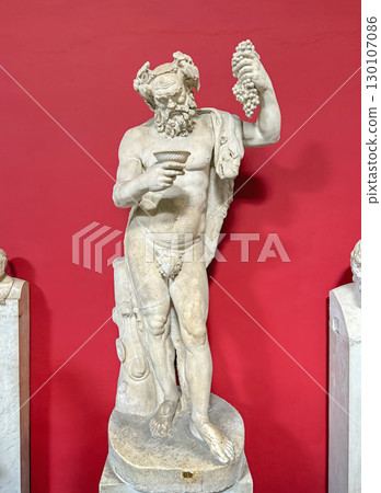 VATICAN CITY, ROME, ITALY - AUGUST 02, 2025 A full-body marble statue of Silenus holding a bunch of grapes and a cup of wine, displayed on a pedestal inside Pio-Clementino museum VATICAN CITY, ROME, ITALY - AUGUST 02, 2025 A full-body marble statue of Silenus holding a bunch of grapes and a cup of wine, displayed on a pedestal inside Pio-Clementino museum 130107086