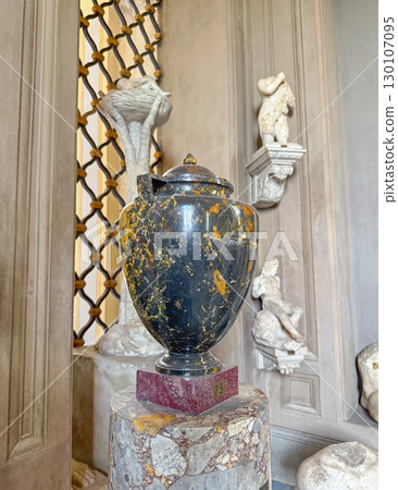 A striking black and gold speckled marble urn sits on a red marble base, set against a wall with various Greek or Roman classical sculptures in the background 130107095