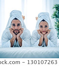 A young adult man and woman, both with white towels on their heads and matching bathrobes, lie smiling on a white bed. Generative AI. 130107263