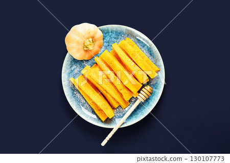 Baked pumpkin slices with cinnamon and honey on a blue plate on a deep blue background. Vegan dessert, healthy food. Autumn food and comfort meal. Flat lay Baked pumpkin slices with cinnamon and honey on a blue plate on a deep blue background. Vegan dessert, healthy food. Autumn food and comfort meal. Flat lay 130107773