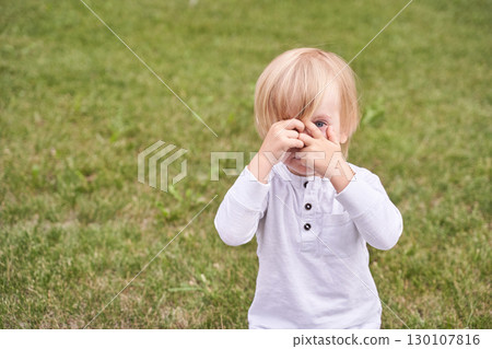 Blonde caucasian toddler playing peekaboo in park wearing white long sleeve Blonde caucasian toddler playing peekaboo in park wearing white long sleeve 130107816
