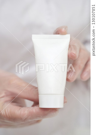 Gloved hands holding white tube of skincare product for beauty and healthcare 130107851