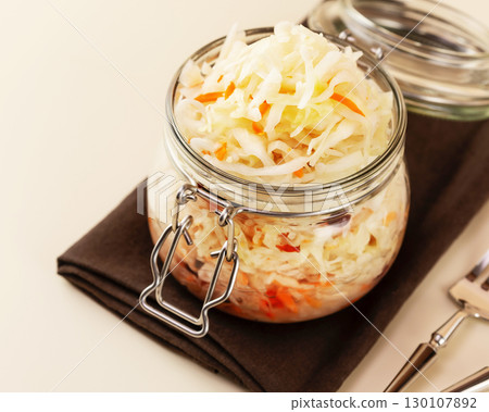 Probiotic food. Pickled or fermented vegetables. Sauerkraut in glass jar on a brown napkin with fork and knife on a kitchen table. Home food preserving or canning. Close up Probiotic food. Pickled or fermented vegetables. Sauerkraut in glass jar on a brown napkin with fork and knife on a kitchen table. Home food preserving or canning. Close up 130107892