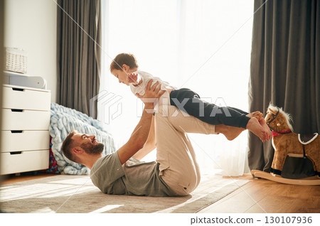 Father is lying down, holding boy in hands. Man is playing with his little son in the domestic room 130107936