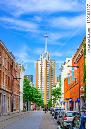 High residential building with spire and narrow street with parking cars in Leuven city center in sunny summer day, blue sky background, Flemish Region, Flemish Brabant province, Belgium 130108297