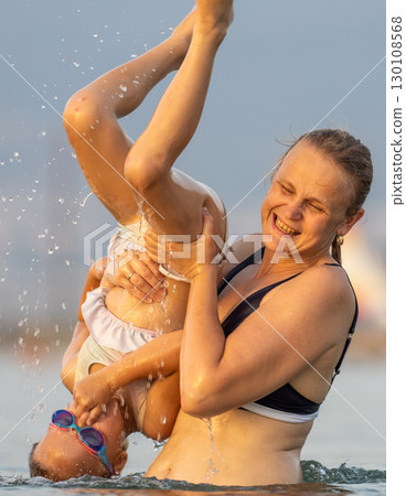 Happy mother holding her daughter upside down while laughing together, enjoying playful moments in the sea during a sunny summer day Happy mother holding her daughter upside down while laughing together, enjoying playful moments in the sea during a sunny summer day 130108568