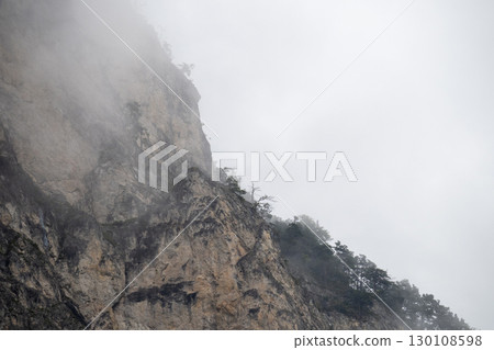 Misty mountain slope shrouding rugged terrain, rocky landscape veiled by low hanging clouds, minimal greenery emerging Misty mountain slope shrouding rugged terrain, rocky landscape veiled by low hanging clouds, minimal greenery emerging 130108598