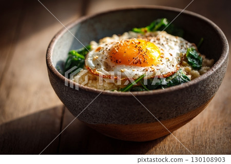 A bowl of creamy oatmeal topped with a fried egg and cooked spinach on a wooden table, in warm sunlight. Healthy breakfast concept 130108903